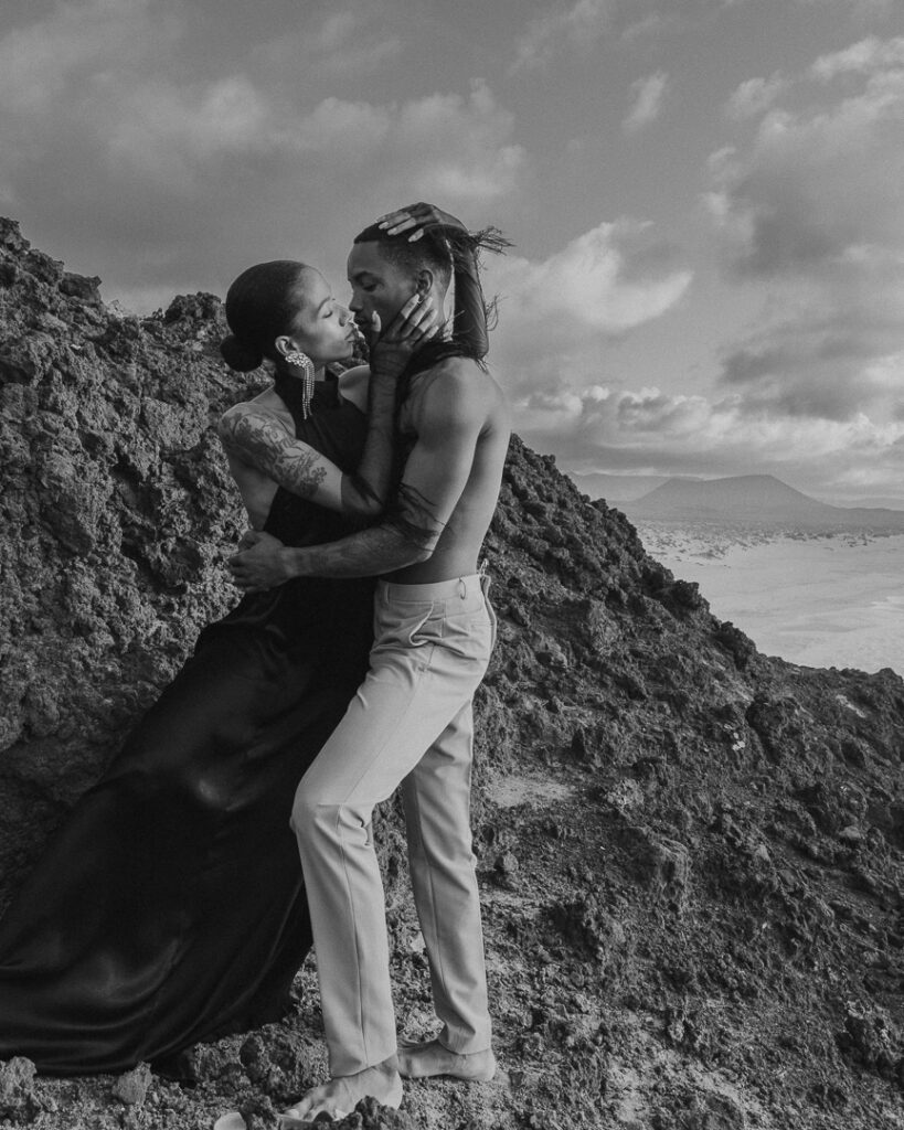Young couple pose and kiss on a cliff overlooking ocean at Playa de las Conchas. 