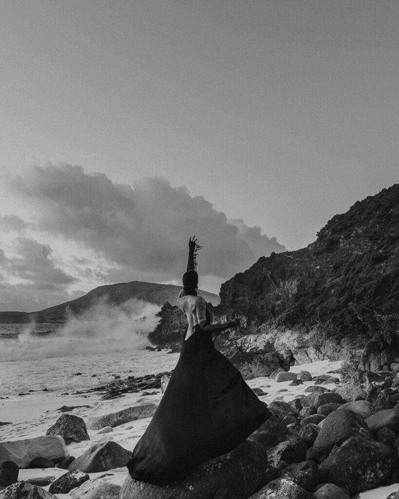 Woman in beatiful black dress surrounded by rocks and the ocean waves on a beach at Playa de las Conchas.